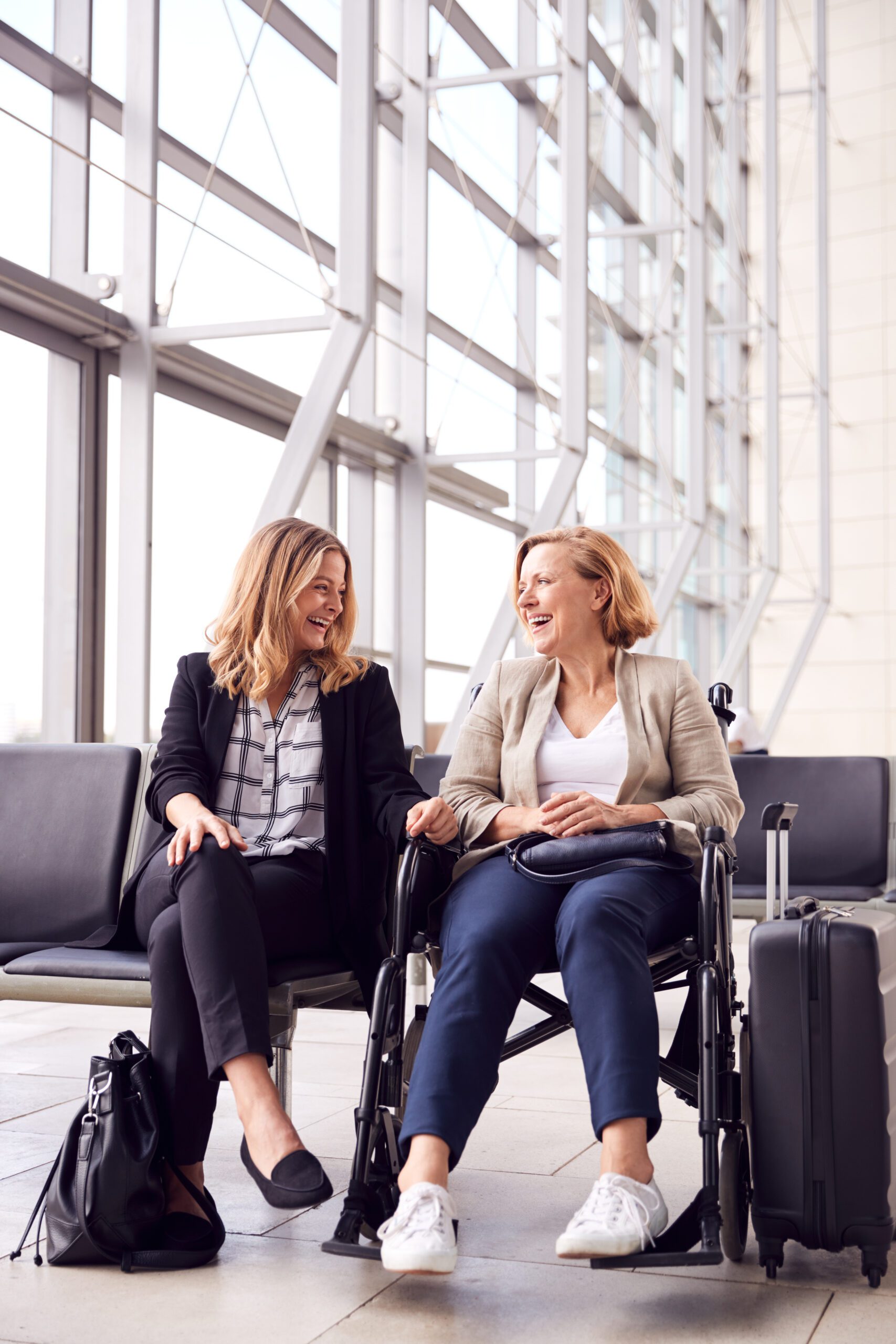 Businesswoman Sitting In Airport Departure With Female Colleague In Wheelchair Talking Together